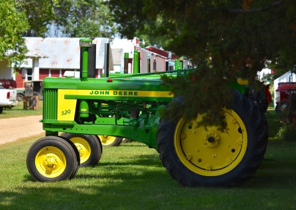 A classic John Deere tractor parked on a sunny farm surrounded by greenery and rural buildings.