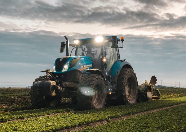 Big tractor with headlights and plow riding on green agricultural plantation in countryside on farmland against cloudy sky during harvest season